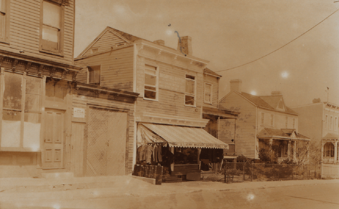 historic photo of wood frame houses, one with a wide awning and signage for a military supply store