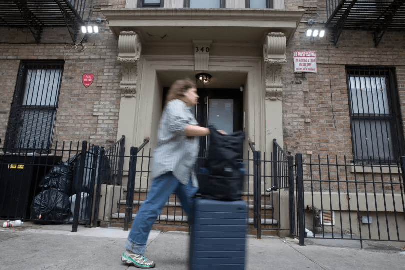 co-living- A young person with an “Away” suitcase walks by Outpost Club tenement building in Bed Stuy