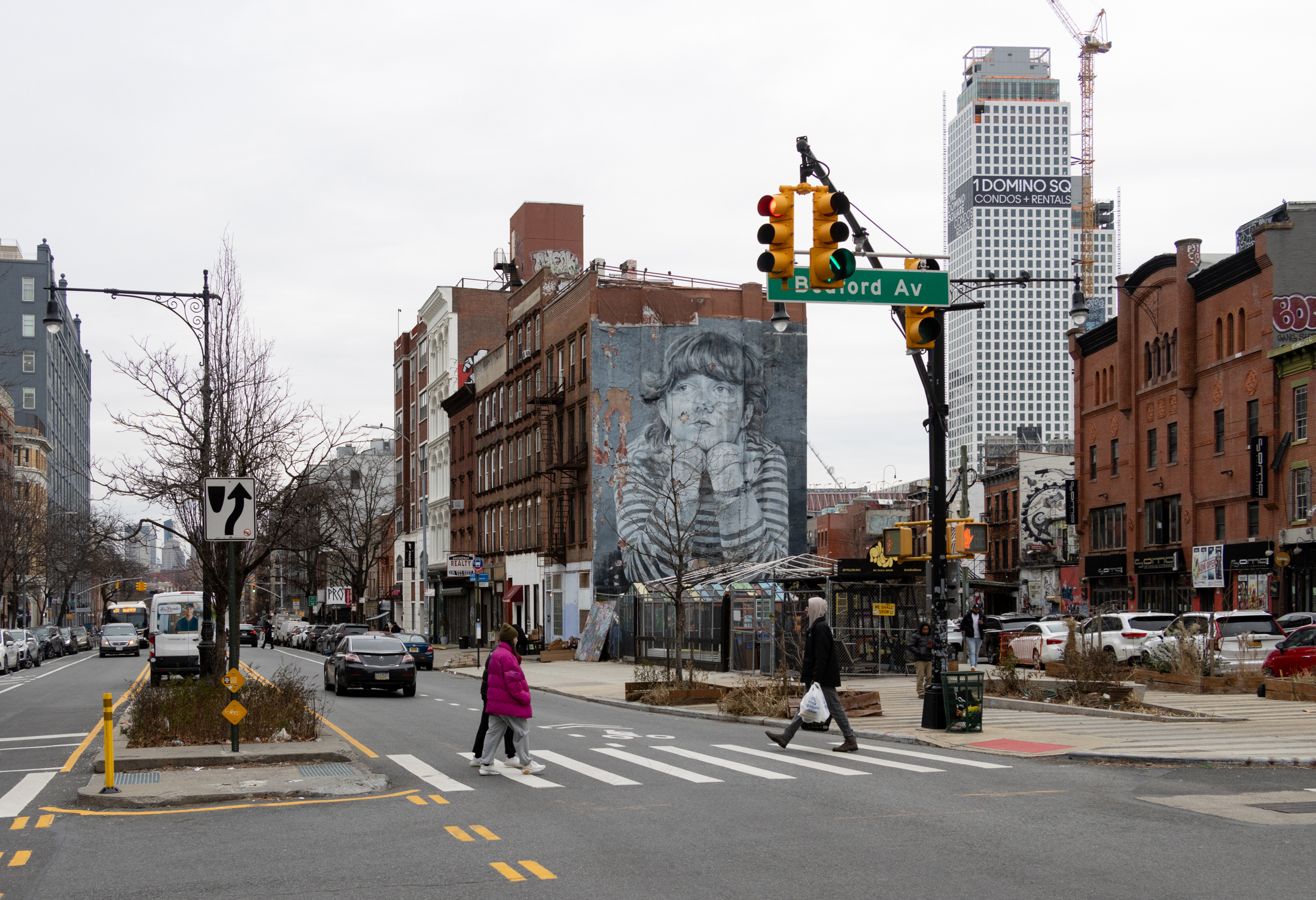 people crossing broadway with view of triangular site and 1 domino tower in the background