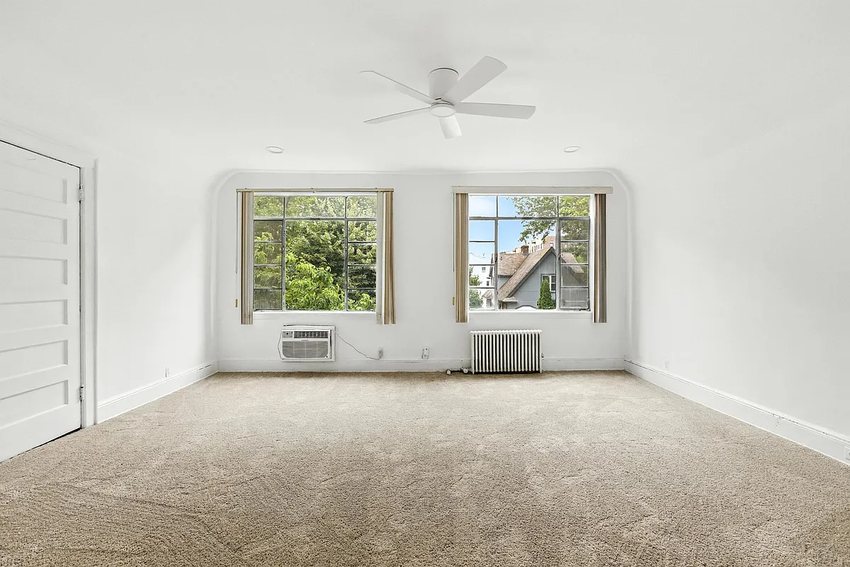 a bedroom with beige carpet, two windows, white walls and a ceiling fan