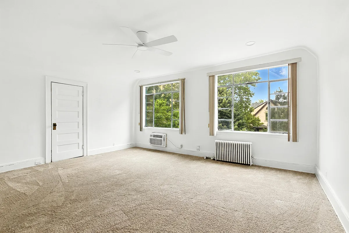 a bedroom with beige carpet, two windows, white walls, and a ceiling fan