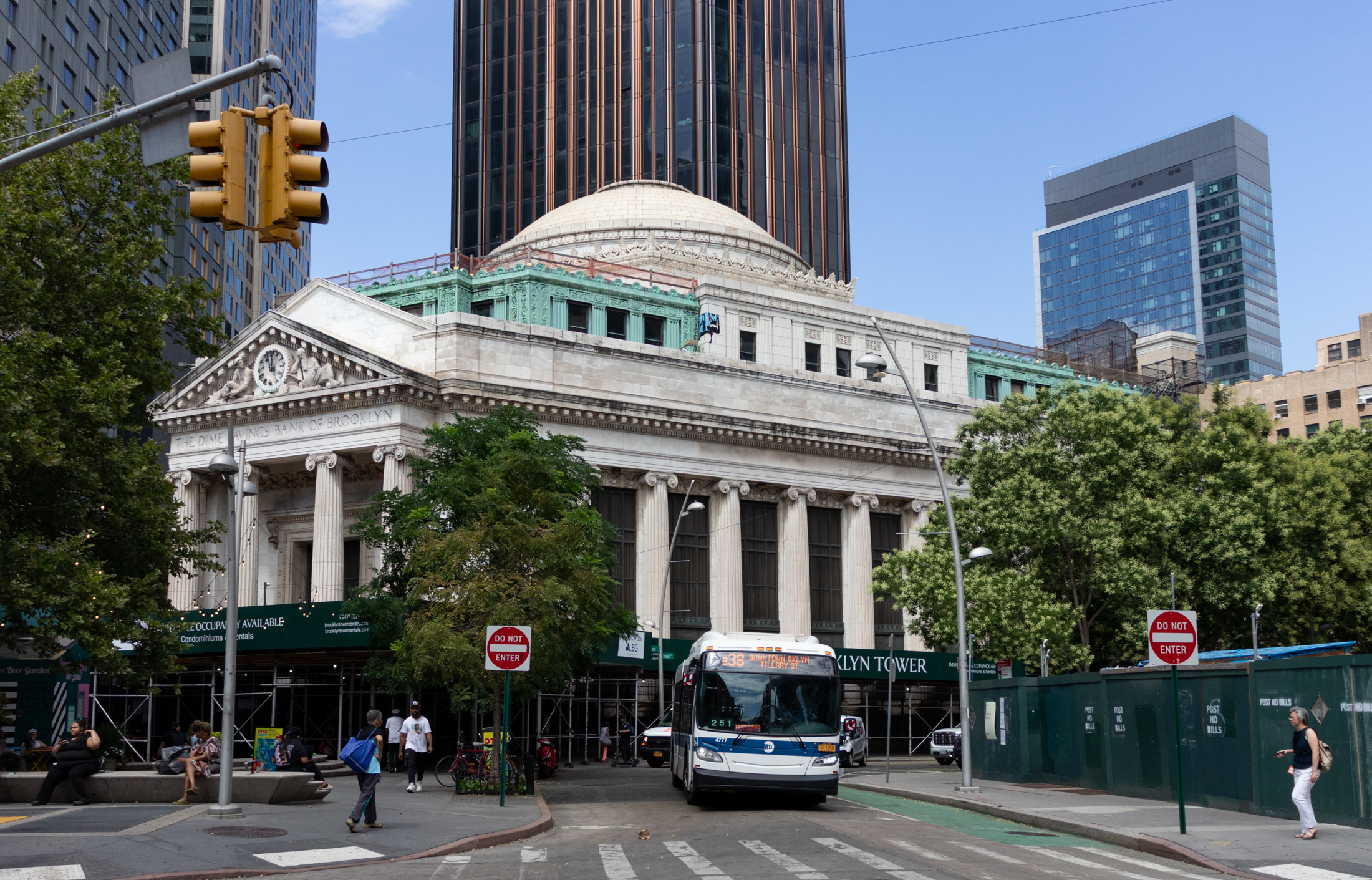 a b38 bus nearing fulton street in front of the dimes saving bank which has a green construction fence